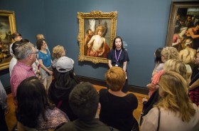 Tour guide stands in front of crowd of visitors. Behind the guide is a painting of an angel.