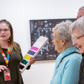 An AGO Art Educator stands to the left holding a board with painted colours. They wear a green shirt and a rainbow lanyard. On the right, there are three elderly individuals smiling. A painting can be seen in the background out of focus. 