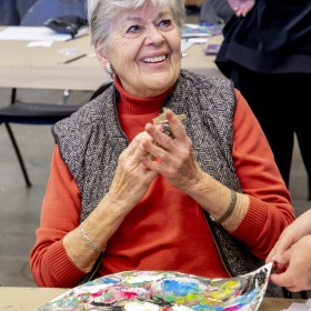 An individual in an orange shirt and grey vest sits at a table in front of an abstract painting. They smile, clasping their hands together. A figure in black, with their back turned to the camera, can be seen in the background with their hand of their hips.  