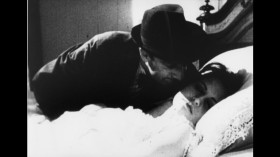 Black and white film still of a man leaning over a woman lying in bed from Betty Ferguson's Kisses