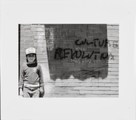 Black and White photograph of a young boy wearing a baseball cap standing against a brick wall with the words "culture Revolution" written