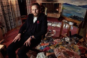 Portrait of artist Ragnar Kjartansson wearing a black suit, sitting in a chair surrounded by oil paint palettes and a painting on an easel