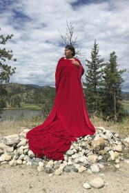 Photo of artist Lori Blondeau standing atop a pile of rocks and stones, lake and hill in the background. She is wrapped in a voluminous swath of deep red fabric that she holds at her chest and pools at her feet. 