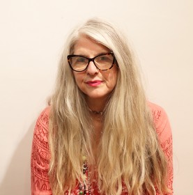 Headshot of a woman with long, straight grey hair, black framed glasses and a pink top