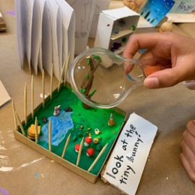 A hand holding a magnifying class over a miniature plasticine sculpture of a garden