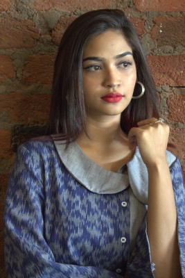 Image of Nuzhat. She wears bright red lipstick, hoop earings and a blue collared shirt. Her arm is pointed upwards with her hand clenched in a fist. She looks away from the camera.