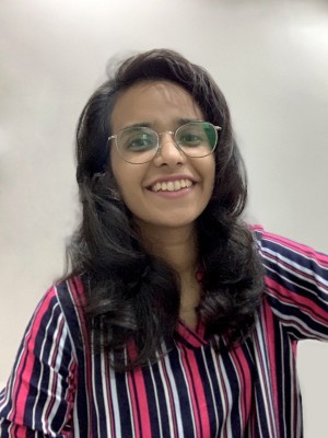 Shivani is pictured in this photo. She smiles, staring into the camera. She wears a vertical stripe shirt with pink, blue and white lines. She is pictured with long brown curly hair and thin wireframe glasses.