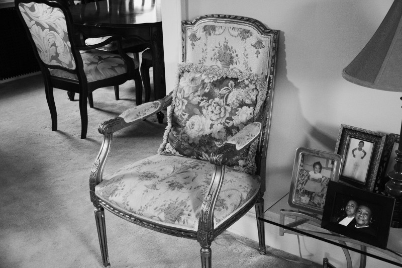 black and white image of a upholstered chair beside a table with framed family photos