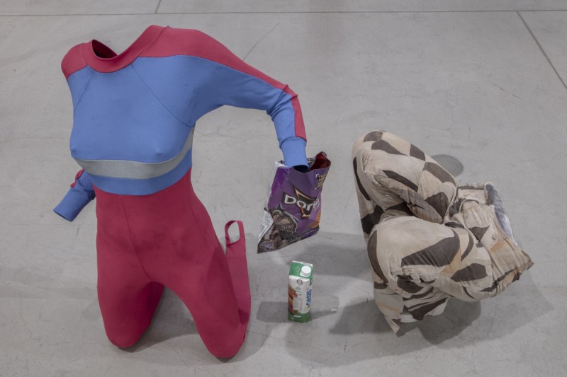 installation photo of sculpture featuring woman's leotard, doritos chips bag, beverage tetra pack and patchwork pants. 