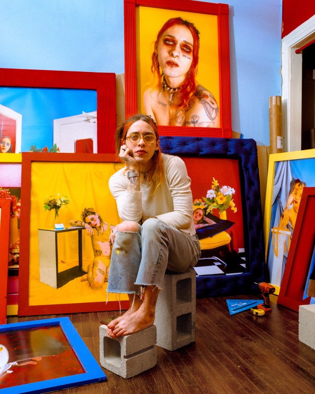 image of artist sitting on cinder blocks surrounded by their frames self portraits, all in bright primary colours