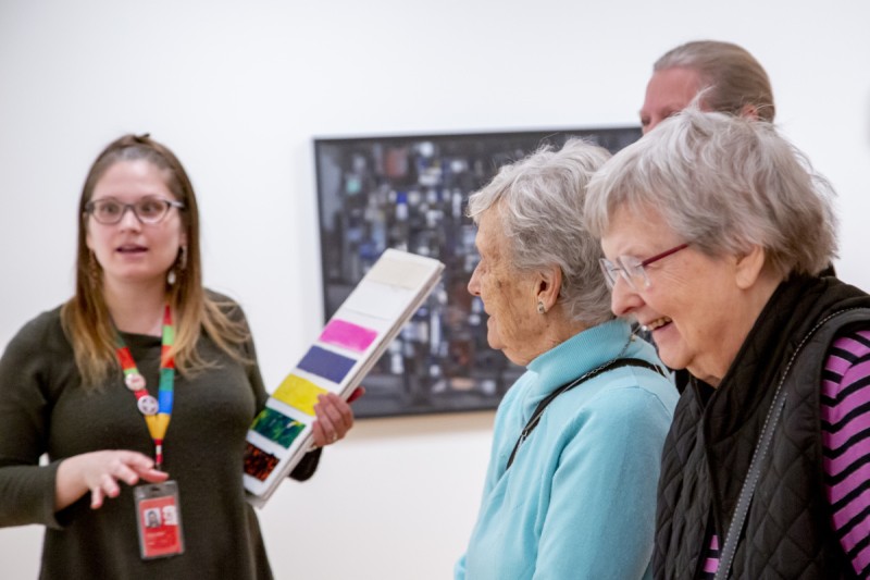 An AGO Art Educator stands to the left holding a board with painted colours. They wear a green shirt and a rainbow lanyard. On the right, there are three elderly individuals smiling. A painting can be seen in the background out of focus. 