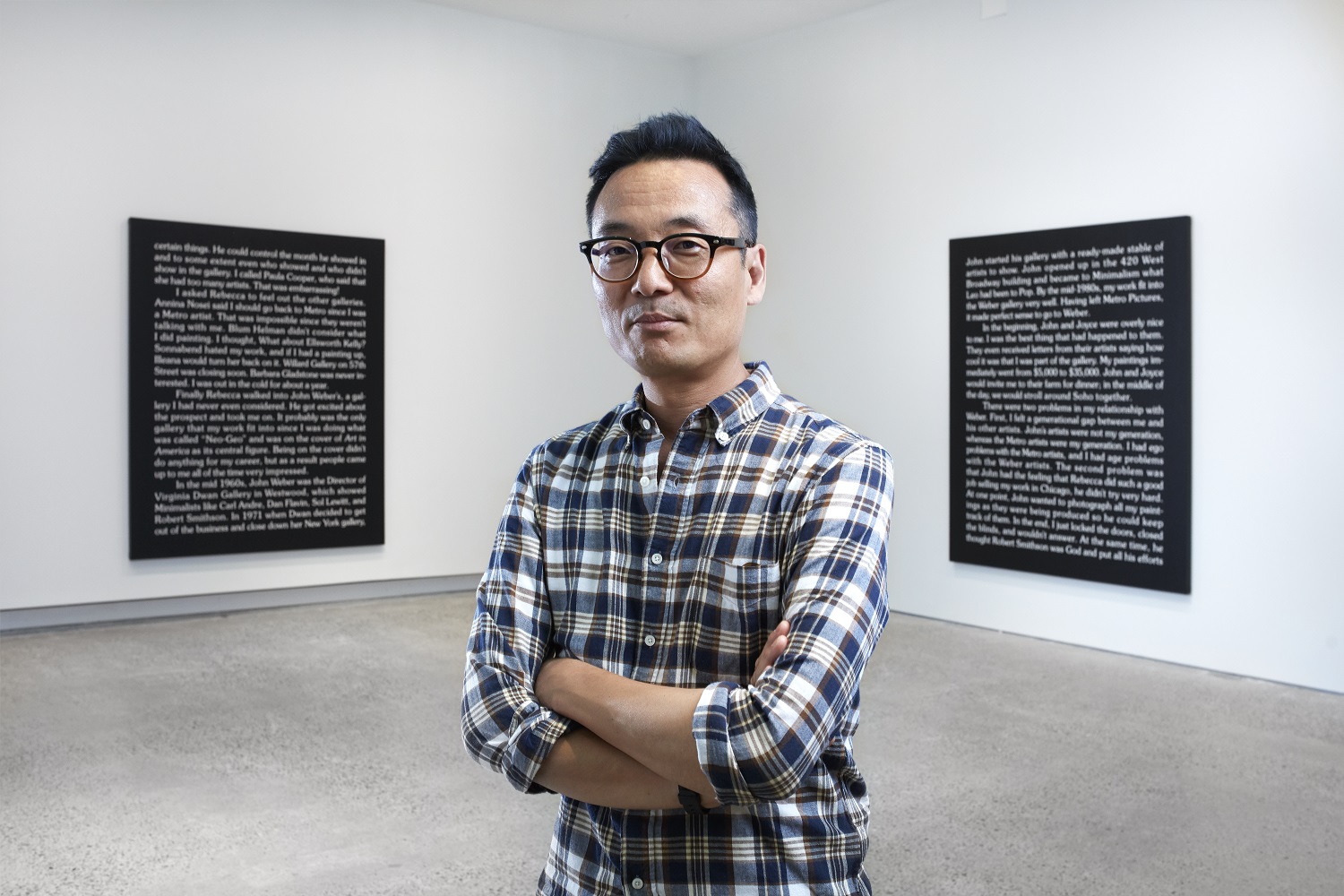 Artist Ron Terada stands in a gallery, in front of two of his paintings with large blocks of white text on a black canvas.