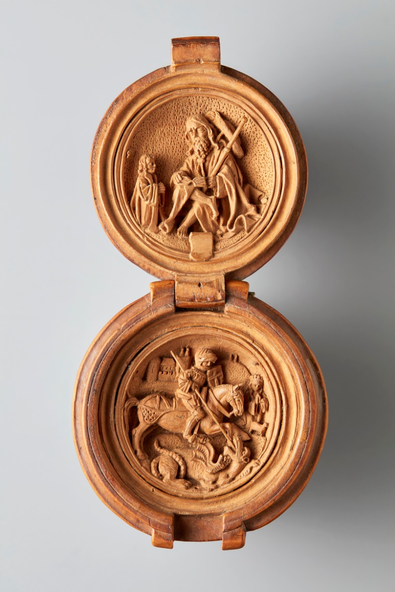 Close up of a small wooden bead, open, depicting intricately carved scenes of Saint James and Saint George