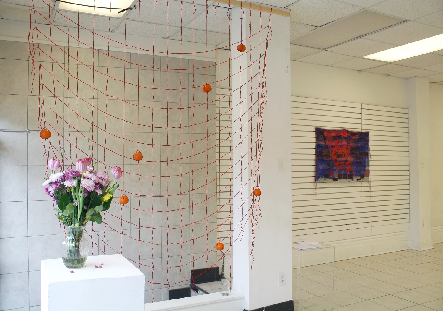 A bouquet of vases, a red grid of string, and a red and blue tapestry hang in a gallery with white walls.