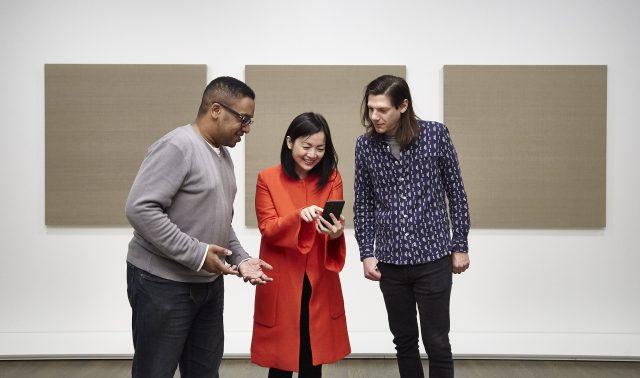 2 men and a woman look at a phone in front of 3 panels of art