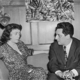 image of artists Joan MItchel and Jean-Paul Riopelle seated on a sofa in front of Riopelle's paintings
