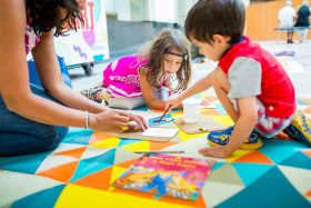Two children on the floor drawing and painting with an adult.