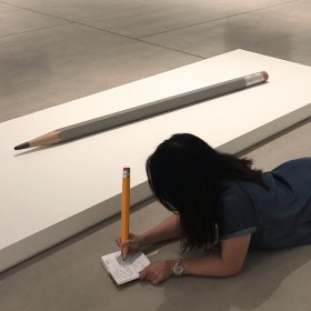 person on floor next to Celmins pencil sculpture, writing with large pencil