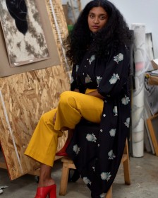 Artist Rajni Perera seated in her studio wearing a long black coat and yellow trousers