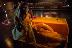 image of performance by Kama La Mackerel reclining wearing a saffron yellow sari with the same textile on a table to their left. in the background circles made of rose petals.