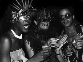 Three figures are pictured in this black and white scene. The figure on the far left wears a metallic costume mask with feathers which protrude upwards. Their mouth is open as they hold a glass in their hand. The figure in the middle wears a dark feather boa, presents a half-smirk and a feather black costumer mask. They are also holding a glass with an opaque liquid. The figure on the far right has short hair and wears a buttoned shirt that has been unbuttoned. They stare into the camera wearing sunglasses.