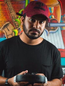 Image of DJ Kevin in a black tshirt, dark red baseball cap standing in front of bright orange and red mural