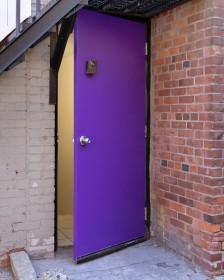 vertical image of the exterior of a deep purple door, slightly ajar to the outside, flanked by brick walls on either side of the door. The bricks on the right are bare, those on the left are painted light beige. 