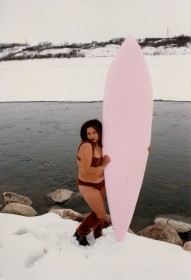 Photography of artist Lori Blondeau as the Lonely Surfer Squaw, standing beside a river on snow-covered ground, wearing a buffalo skin bikini and boots, holding a large pale pink surf board.