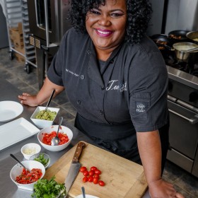 Chef La-toya standing in a kitchen, with a knife, cutting board and vegetables. She is smiling up into the camera. 