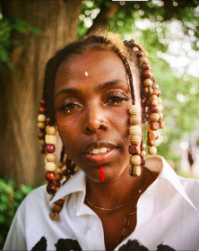 Close up photograph of a person with deadlocks with beads in a white blouse with trees in a blurred background