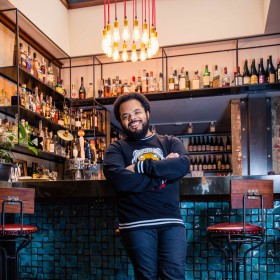 Roger Mooking standing in front of a bar at a restaurant, smiling at the viewer with his arms crossed. 