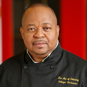 Chef Selwyn Richards wearing a black chef's uniform, looking at the viewer, with a blurred red and black background behind. 