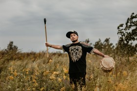Beny standing outside in a field, holding traditional instruments in their outstretched hands, one in each hand.