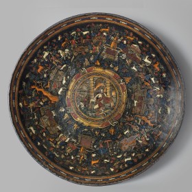 image of a Mexican tray (batea) from 1650. The tray is a circle with a black background and small illustrations of animals, trees, fountain in yellows, whites, reds, and greens