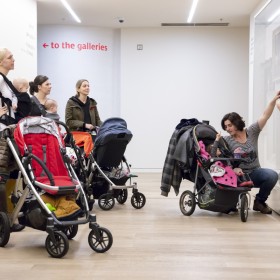 A group of parents carrying babies behind strollers and looking at art