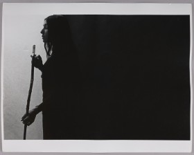 Greyscale photograph of a woman's profile standing against a black block and carrying a long stick