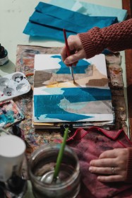 A hand holding a paintbrush and painting over a collage of blue and grey tissue paper in a sketchbook