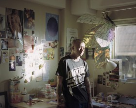 image of a person with a bald head, black tshirt with white text, standing in a room's corner, walls covered in photos, prints and notes, with sunlight streaming through the window