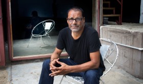 colour photo of artist Christopher Cozier sitting in a white-painted iron patio chair, in front of a mirrored door. The mirrored door's reflection shows the chair but not Cozier. 