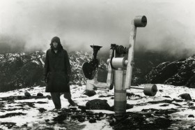 black and white photo of artist Michael Snow in a wintry, hilly landscape, bundled in hat and parka, striding beside a metal piece