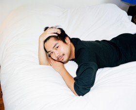 Photograph of a man laying on a large white pillow. The man has one hand on his head and is wearing a black shirt.