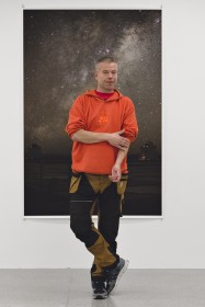 photo of wolfgang tillmans in an oragen hoodie standing in front of his photo of the milky way