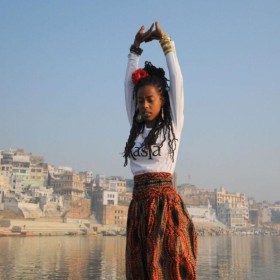 image of Donisha Prendergast standing with her arms stretched overhead with a coast line behind her