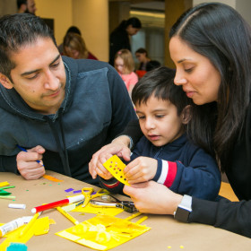 Parents helping a young child sitting at a table making a craft with markers, paper and scissors.