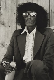 Black and white photo of a person with long hair, reflective sunglasses, frilly shirt and suit holding a plastic juice bottle staring at the camera