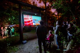 A screen in a dark wooded area displays a video game, as people stand and play and watch