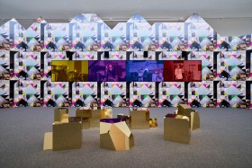 A photograph of Sonia Boyce's artwork. Polygonal, golden sculptures are on the ground, in front of a wall panels in a kaleidoscope-style pattern, with 4 centre coloured panels, with tinted photographs of four people.  