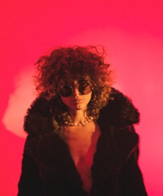 Kiesza wearing sunglasses and faux fur in front of pink background