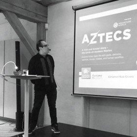 A black and white photo of Fransisco Guevara at a podium, giving a lecture. They are wearing a dress shirt, pants, and sneakers. There is a presentation to his left, where a stylized version of the word "Aztecs" appears. 