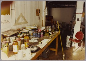 image shows an an artist's studio with a table covered in jars of paint, paper, canvas, and other art supplies, as well as an overflowing ashtray